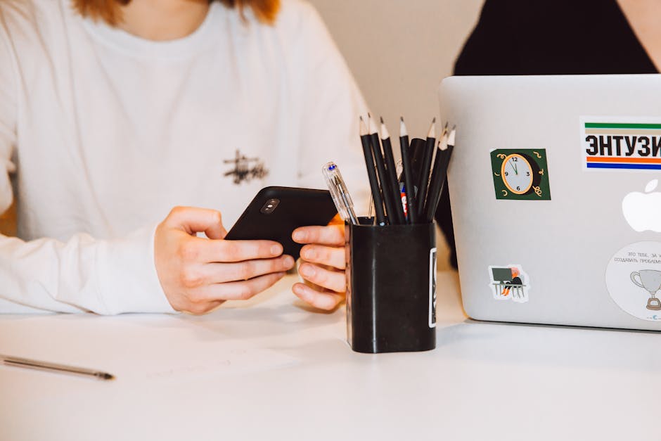 Young adult at a desk texting on smartphone, next to a laptop and stationery holder.