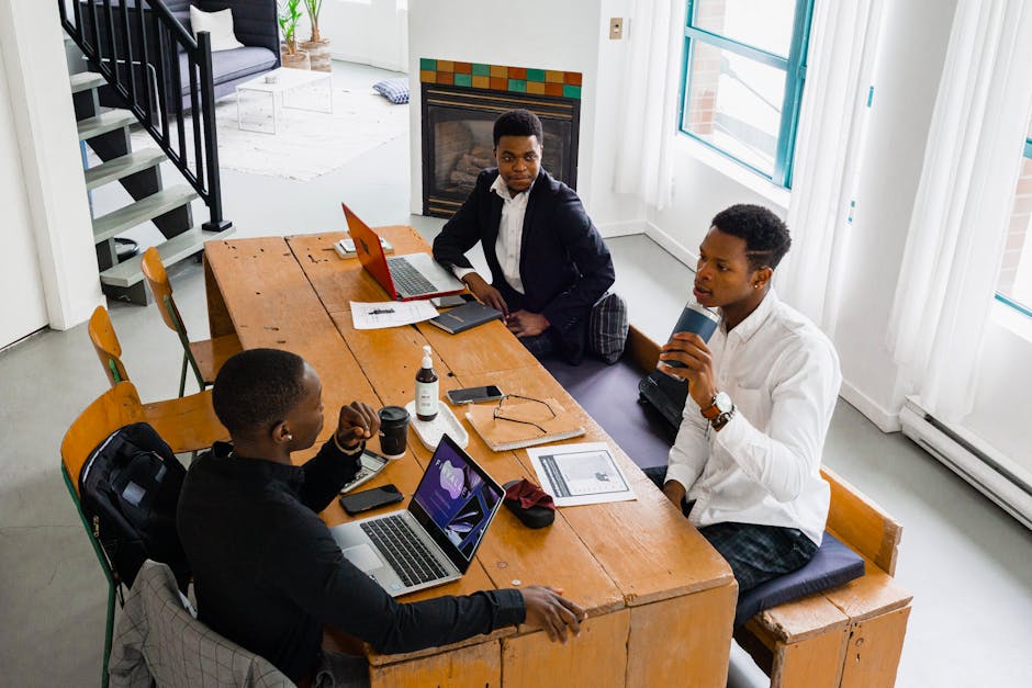 Three colleagues brainstorm indoors using laptops and gadgets at a wooden table.